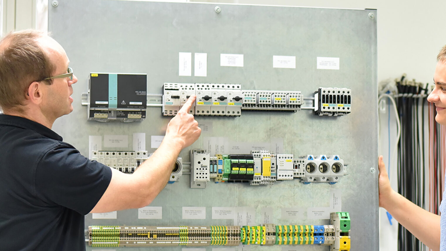Lehrmeister und Auszubildender vor elektrotechnischer Anlage.