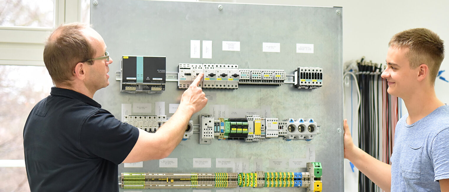 Lehrmeister und Auszubildender vor elektrotechnischer Anlage.